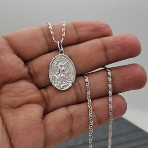 Puede incluir: Un collar con colgante ovalado plateado con una imagen grabada de un santo. El colgante cuelga de una delicada cadena. La imagen representa a un santo con alas y una espada. El collar se muestra sobre un fondo neutro.