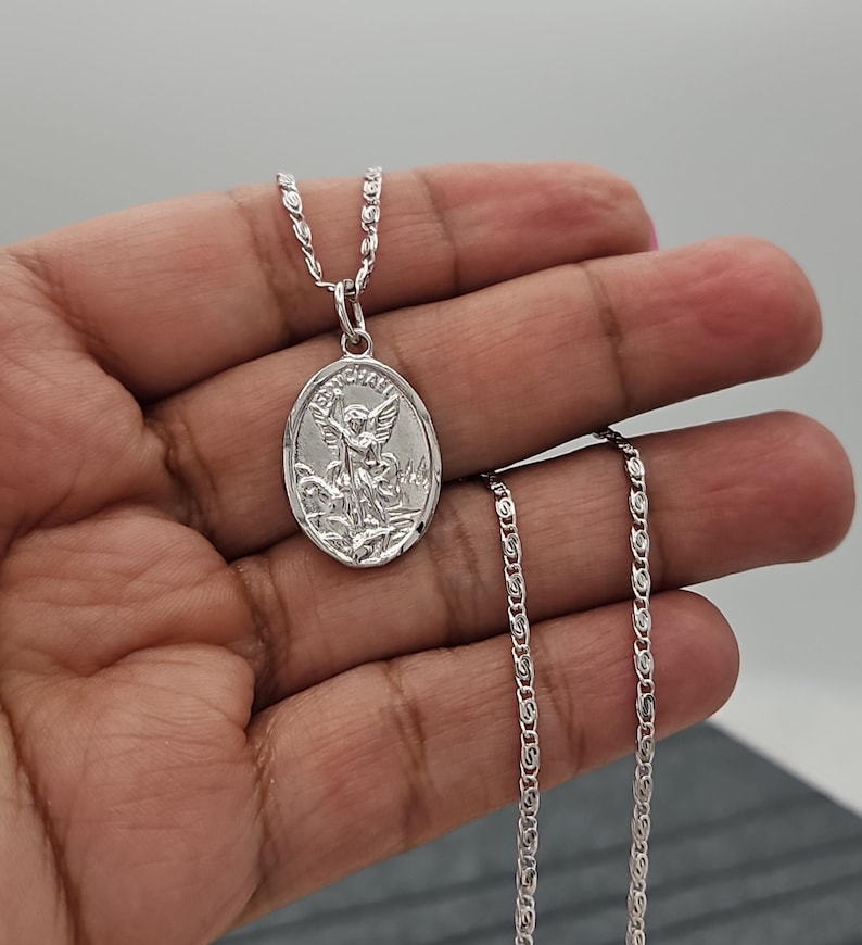 Puede incluir: Un collar con colgante ovalado plateado con una imagen grabada de un santo. El colgante cuelga de una delicada cadena. La imagen representa a un santo con alas y una espada. El collar se muestra sobre un fondo neutro.