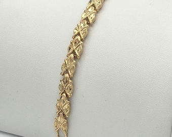 Op de afbeelding: Een goudkleurige armband bestaande uit een herhalende reeks onderling verbonden, getextureerde bladvormige schakels. Het metalen oppervlak heeft een gepolijste afwerking met ingewikkelde, geëtste details op elk segment, wat een doorlopend, decoratief kettingontwerp vormt.