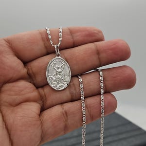 Puede incluir: Un collar con colgante ovalado plateado con una imagen grabada de un &aacute;ngel. El colgante cuelga de una delicada cadena. El collar se muestra sobre un fondo neutro.
