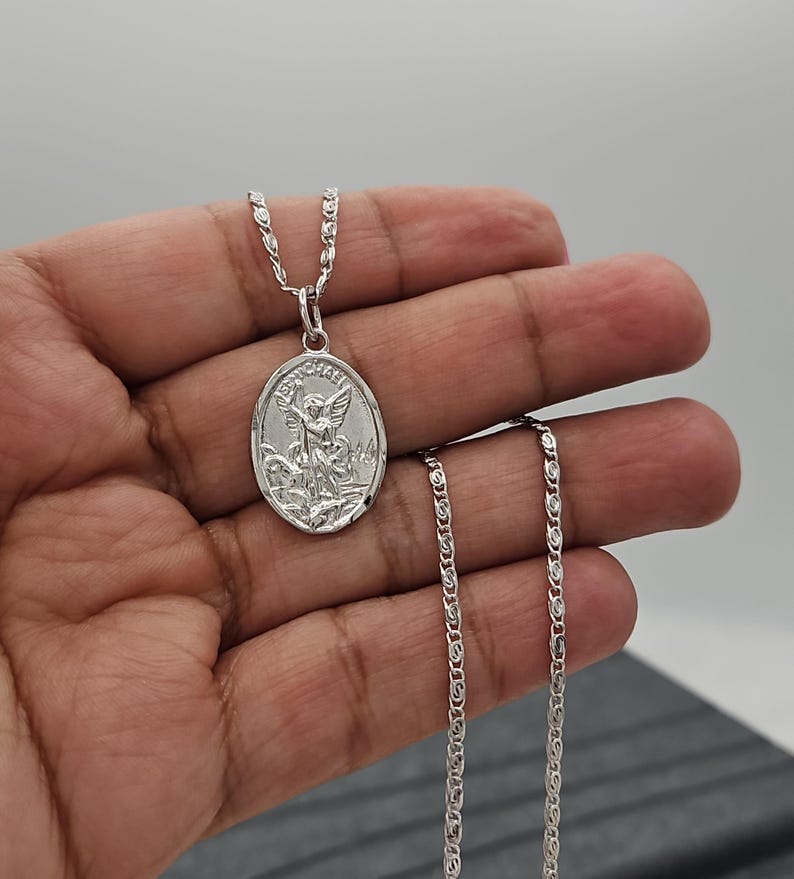 Puede incluir: Un collar con colgante ovalado plateado con una imagen grabada de un &aacute;ngel. El colgante cuelga de una delicada cadena. El collar se muestra sobre un fondo neutro.