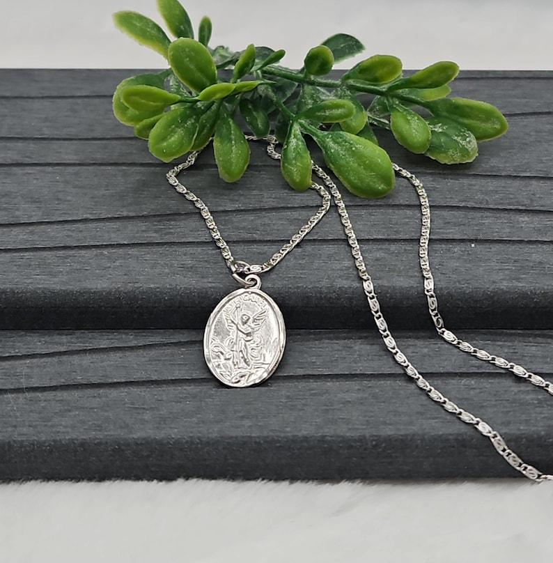 Puede incluir: Un collar plateado con un colgante ovalado detallado que presenta un dise&ntilde;o de &aacute;ngel. La cadena es delicada y descansa sobre una superficie gris oscuro, con hojas verdes decorativas en el fondo.