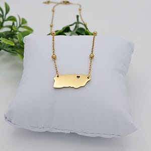 May include: Gold necklace featuring a pendant shaped like the outline of Puerto Rico with a small heart cutout. The necklace has a delicate chain with small round beads.