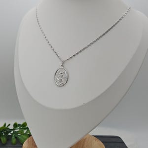 Puede incluir: Un collar plateado con un colgante que representa a un santo. El colgante ovalado cuelga de una fina cadena. El collar se exhibe en un maniqu&iacute; blanco. El colgante es probablemente de plata.