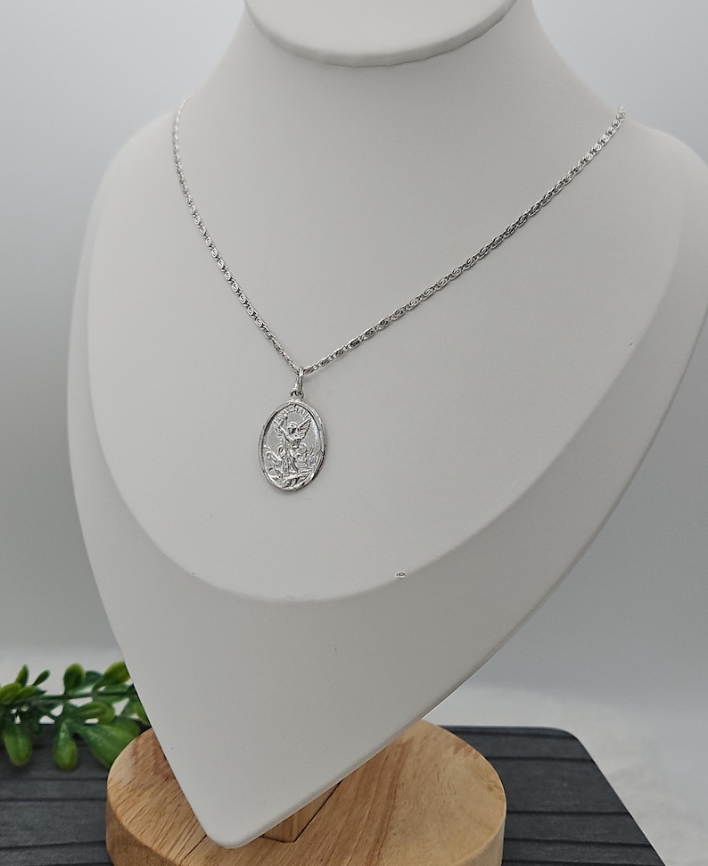 Puede incluir: Un collar plateado con un colgante que representa a un santo. El colgante ovalado cuelga de una fina cadena. El collar se exhibe en un maniqu&iacute; blanco. El colgante es probablemente de plata.