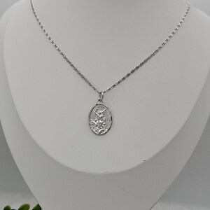 Puede incluir: Un collar plateado con un colgante ovalado que representa una figura grabada. El colgante cuelga de una fina cadena. El collar se exhibe en un maniqu&iacute; blanco.