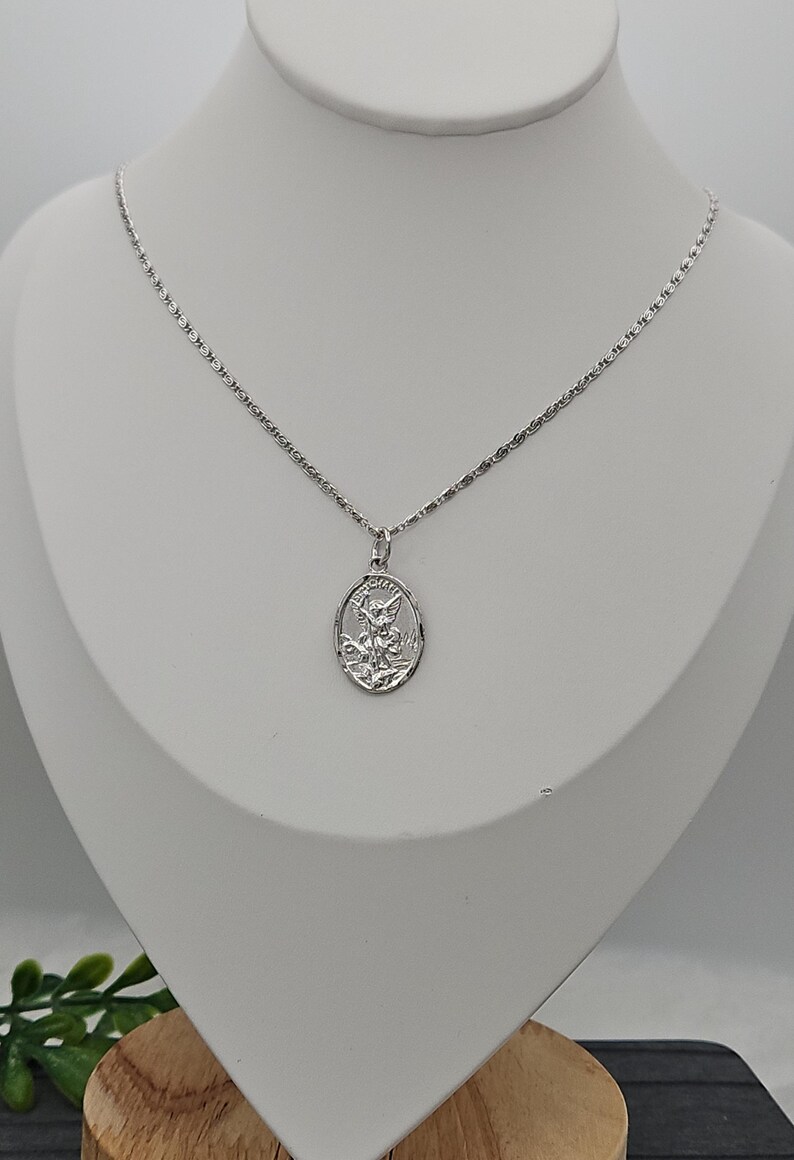 Puede incluir: Un collar plateado con un colgante ovalado que representa una figura grabada. El colgante cuelga de una fina cadena. El collar se exhibe en un maniqu&iacute; blanco.