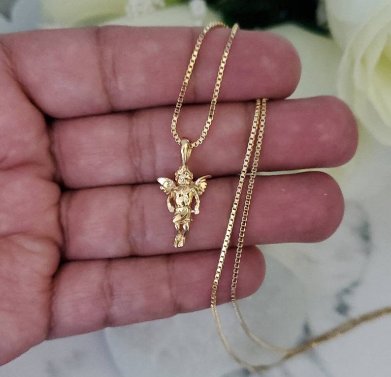 Puede incluir: Un collar de cadena de oro con un peque&ntilde;o colgante de querub&iacute;n. La cadena es de estilo cadena de caja.