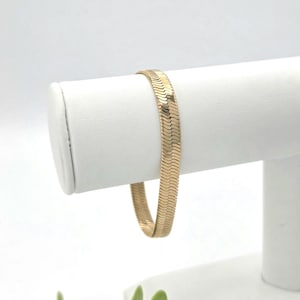 Könnte beinhalten: Ein goldfarbenes Fischgräten-Kettenarmband mit einem flachen, flexiblen und eng gewebten Gliedermuster. Das Armband hat eine polierte, reflektierende Oberfläche, die das Licht einfängt und ein durchgehendes, einheitliches Design über die gesamte Länge zeigt.
