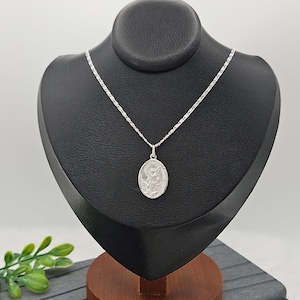 Puede incluir: Un collar con colgante plateado exhibido sobre un busto negro. El colgante ovalado presenta un dise&ntilde;o en relieve detallado. La delicada cadena plateada cuelga del busto, que est&aacute; sobre una base de madera.