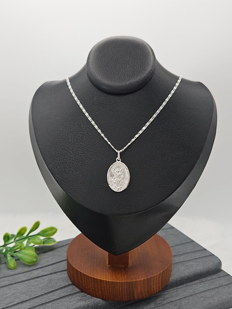 Puede incluir: Un collar con colgante plateado exhibido sobre un busto negro. El colgante ovalado presenta un dise&ntilde;o en relieve detallado. La delicada cadena plateada cuelga del busto, que est&aacute; sobre una base de madera.