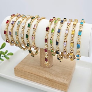 May include: A display of gold tone bracelets with colorful square-shaped gemstones. The bracelets are arranged on a wooden stand.