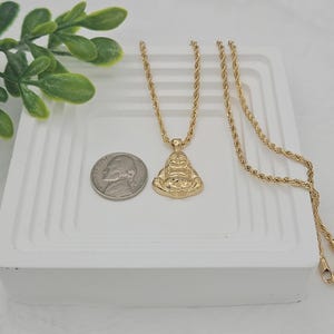 May include: A gold-toned rope chain necklace featuring a pendant shaped like a seated Buddha figure. The pendant is positioned next to a United States nickel coin for scale, showing the relative size of the jewelry piece against the metal currency.