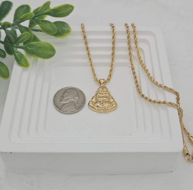 May include: A gold-toned rope chain necklace featuring a pendant shaped like a seated Buddha figure. The pendant is positioned next to a United States nickel coin for scale, showing the relative size of the jewelry piece against the metal currency.