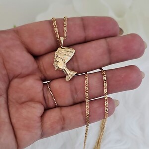 May include: Gold chain necklace with a pendant featuring a profile of an Egyptian queen wearing a headdress.