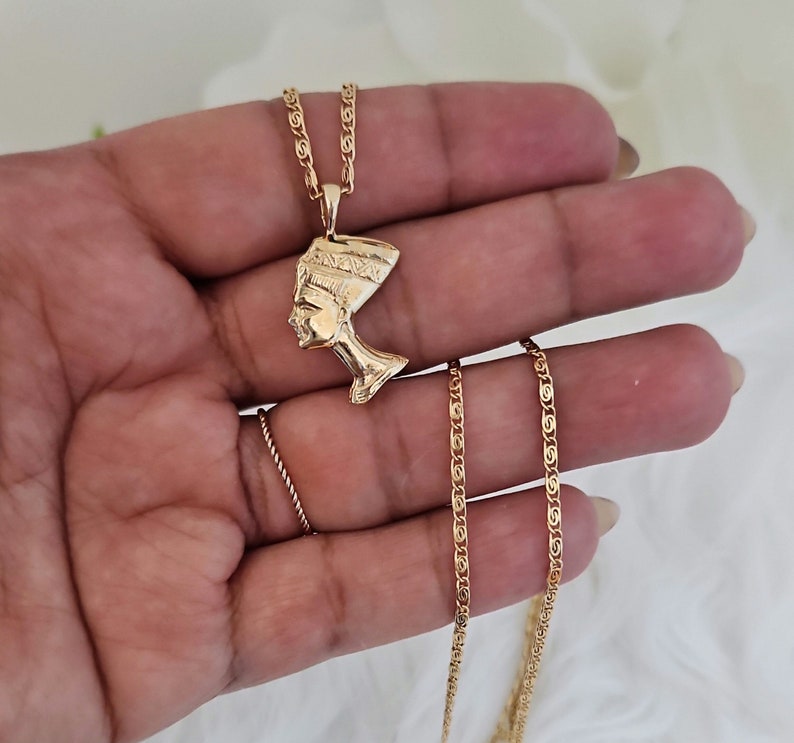May include: Gold chain necklace with a pendant featuring a profile of an Egyptian queen wearing a headdress.