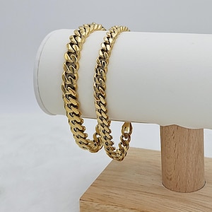 May include: Two gold-tone curb chain bracelets on a wooden display stand. The chains are thick and have a shiny finish.