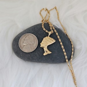May include: Gold chain necklace with a pendant of a woman's profile wearing a headdress. The pendant is on a gray rock with a silver coin.
