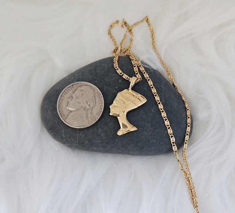 May include: Gold chain necklace with a pendant of a woman's profile wearing a headdress. The pendant is on a gray rock with a silver coin.
