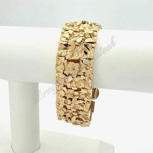 Può includere: Un braccialetto color oro in stile pepita. Il braccialetto è composto da più maglie rettangolari con una superficie strutturata e irregolare. Il braccialetto è esposto su un supporto bianco su sfondo bianco.