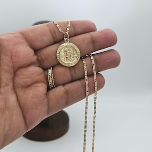 May include: A gold chain necklace with a round pendant featuring a depiction of Saint Christopher. The pendant has the inscription "Saint Christopher Protect Us".