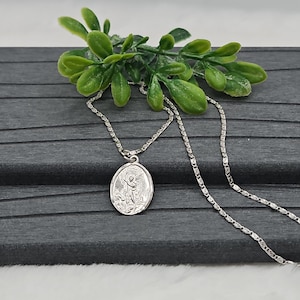 Puede incluir: Un collar con colgante ovalado plateado con un dise&ntilde;o de &aacute;ngel. El colgante cuelga de una delicada cadena. El collar se exhibe sobre una superficie gris oscuro con follaje verde.