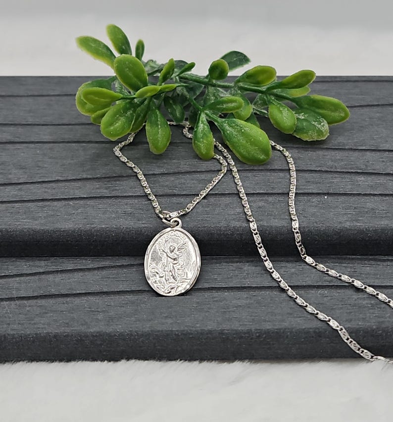 Puede incluir: Un collar con colgante ovalado plateado con un dise&ntilde;o de &aacute;ngel. El colgante cuelga de una delicada cadena. El collar se exhibe sobre una superficie gris oscuro con follaje verde.