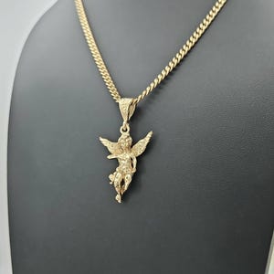 K&ouml;nnte beinhalten: Eine goldfarbene Halskette mit Anh&auml;nger, die ein detailliertes Cherub-Design aufweist. Der Cherub hat ausgebreitete Fl&uuml;gel und h&auml;ngt an einer Panzerkette. Die Halskette wird vor einem schwarzen Hintergrund pr&auml;sentiert.