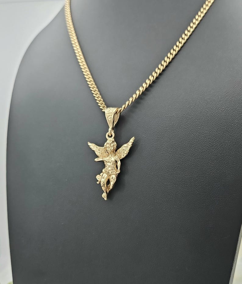 K&ouml;nnte beinhalten: Eine goldfarbene Halskette mit Anh&auml;nger, die ein detailliertes Cherub-Design aufweist. Der Cherub hat ausgebreitete Fl&uuml;gel und h&auml;ngt an einer Panzerkette. Die Halskette wird vor einem schwarzen Hintergrund pr&auml;sentiert.