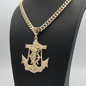May include: A gold-toned metal necklace featuring a thick curb chain and a large, detailed pendant shaped like a ship anchor with a crucifix figure of Jesus Christ centered on the front, displaying a textured, metallic finish throughout the piece.
