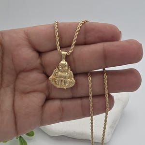 May include: A gold-toned rope chain necklace featuring a pendant shaped like a seated figure with hands resting on the lap. The pendant has a smooth, polished finish and is suspended from a twisted metal chain that drapes down the length of the piece.