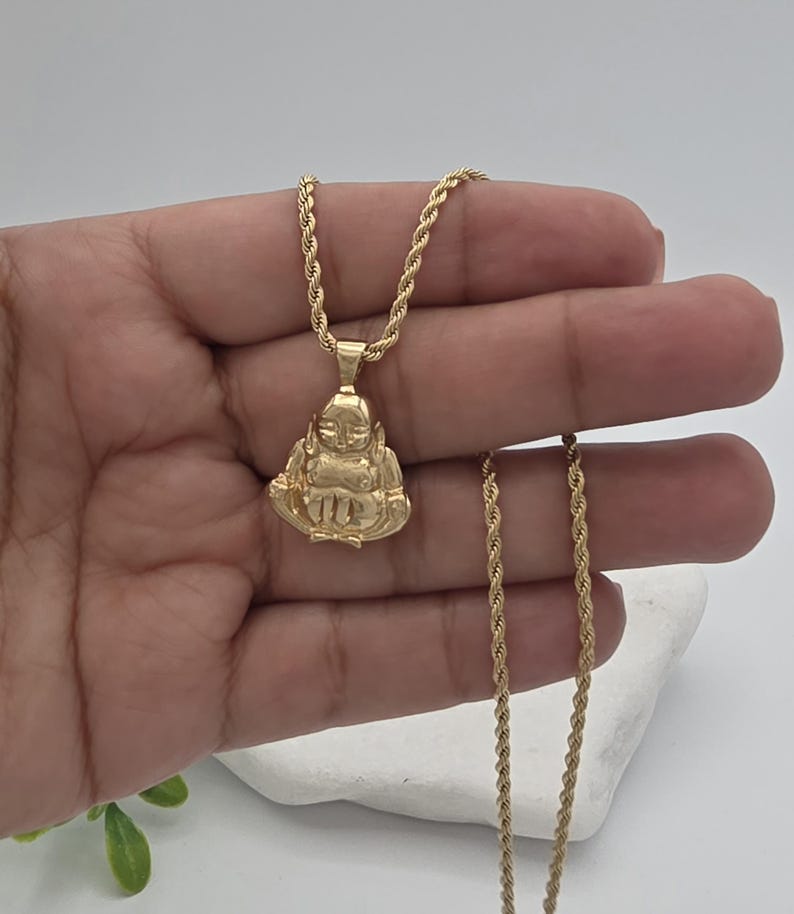 May include: A gold-toned rope chain necklace featuring a pendant shaped like a seated figure with hands resting on the lap. The pendant has a smooth, polished finish and is suspended from a twisted metal chain that drapes down the length of the piece.