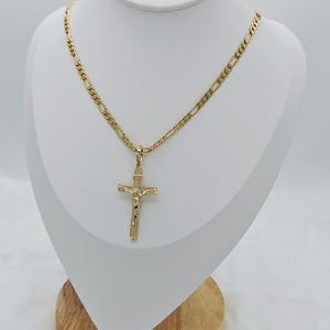 Puede incluir: Un collar de cadena de oro con un colgante de cruz de oro. La cadena es una cadena de estilo figaro.