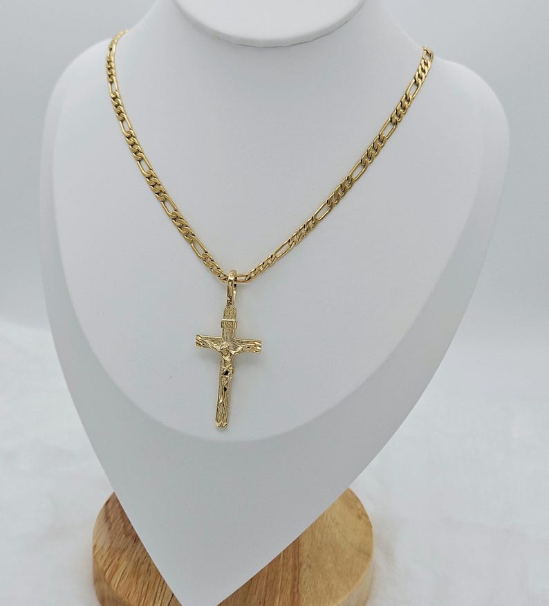Puede incluir: Un collar de cadena de oro con un colgante de cruz de oro. La cadena es una cadena de estilo figaro.