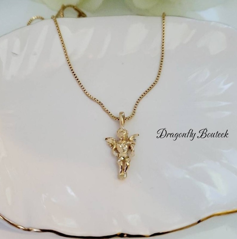 Puede incluir: Un collar de cadena de oro con un peque&ntilde;o colgante de querub&iacute;n de oro. El collar est&aacute; sobre una superficie blanca.