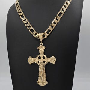 May include: A gold-toned metal necklace featuring a thick, flat curb chain and a large, ornate crucifix pendant. The cross has a textured, nugget-like surface and depicts a central figure of Jesus Christ with a circular halo detail at the intersection.
