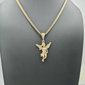 K&ouml;nnte beinhalten: Goldfarbene Halskette mit einem detaillierten Anh&auml;nger eines Engels mit ausgebreiteten Fl&uuml;geln. Der Anh&auml;nger h&auml;ngt an einer Panzerkette. Die Halskette wird vor einem dunklen Hintergrund pr&auml;sentiert.
