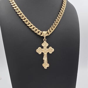 May include: Gold-toned necklace featuring a large cross pendant. The cross has a detailed, textured design, and is suspended from a thick, curb chain. The necklace is displayed on a black stand against a neutral background.