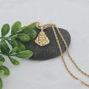 May include: A gold-toned pendant shaped like a seated Buddha figure, suspended from a matching gold-toned rope chain. The pendant features a polished finish and is displayed resting on a smooth, dark gray stone next to green foliage.