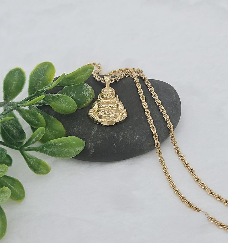 May include: A gold-toned pendant shaped like a seated Buddha figure, suspended from a matching gold-toned rope chain. The pendant features a polished finish and is displayed resting on a smooth, dark gray stone next to green foliage.