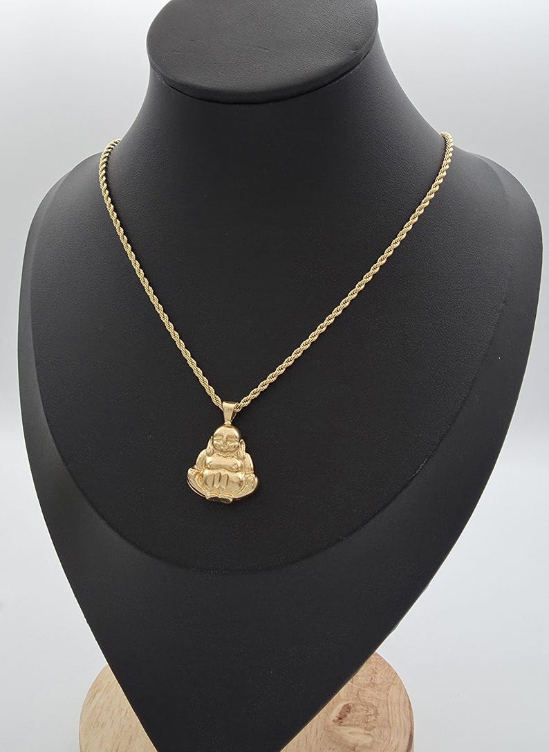 May include: A gold-toned necklace featuring a rope chain and a pendant shaped like a seated, smiling Buddha figure. The pendant has a polished finish and is suspended from a classic twisted metal chain, creating a cohesive gold-colored jewelry piece.