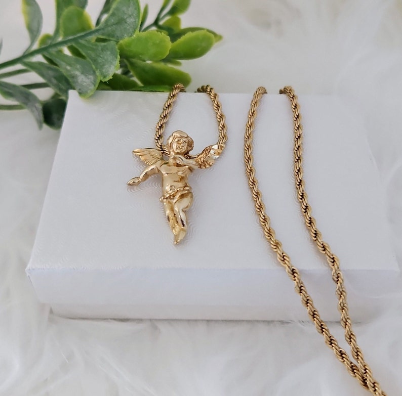 May include: A gold chain necklace with a gold cherub pendant. The chain is a rope style and the pendant is detailed with a cherub holding a flower.