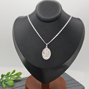 Puede incluir: Un collar con colgante de plata expuesto sobre un busto negro. El collar presenta una cadena delicada y un colgante ovalado con una figura religiosa detallada. El busto est&aacute; sobre una base de madera, con una ramita de hojas verdes en primer plano.