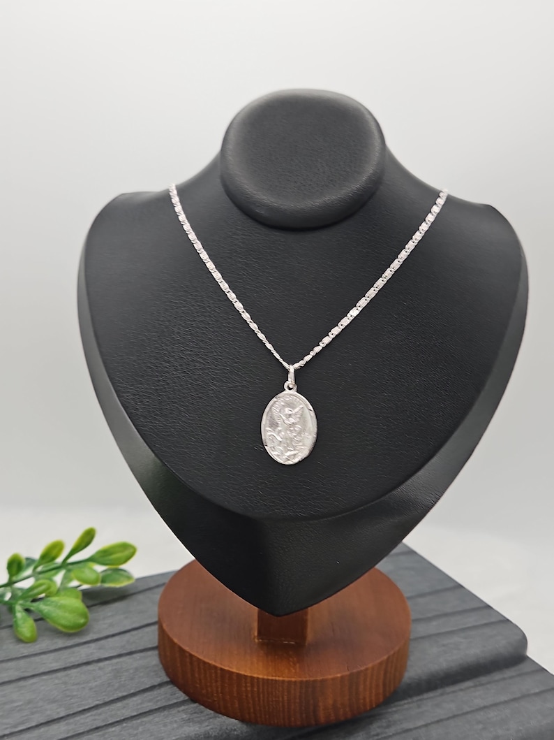 Puede incluir: Un collar con colgante de plata expuesto sobre un busto negro. El collar presenta una cadena delicada y un colgante ovalado con una figura religiosa detallada. El busto est&aacute; sobre una base de madera, con una ramita de hojas verdes en primer plano.