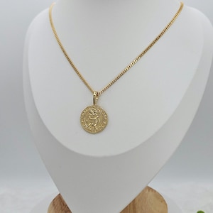 Op de afbeelding: Gouden ketting met een ronde hanger met een afbeelding van Sint Christoffel. De hanger heeft de woorden "Sint Christoffel" en "Bescherm ons" rondom de afbeelding.