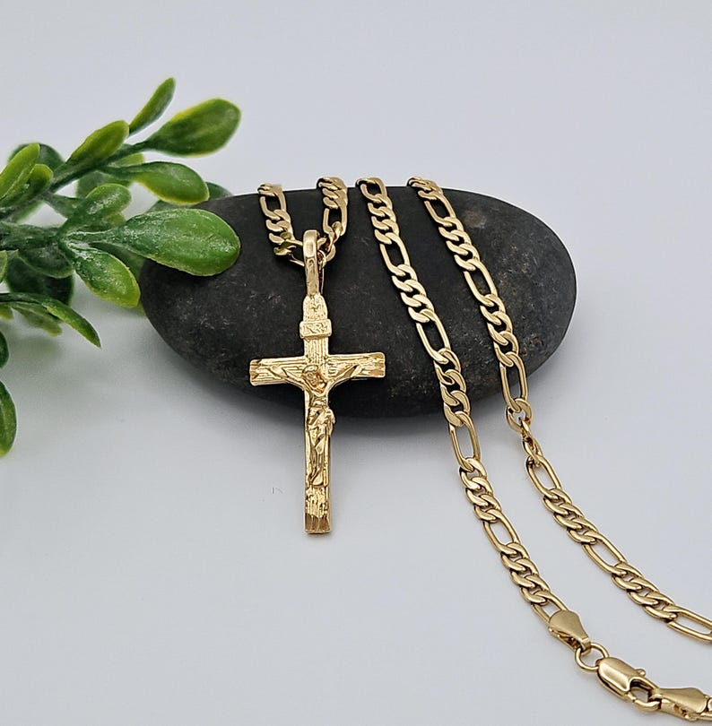 Puede incluir: Un collar de cadena de oro con un colgante de cruz de oro detallado. La cadena es de estilo figaro cl&aacute;sico.