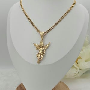 K&ouml;nnte beinhalten: Goldfarbene Halskette mit detailliertem Anh&auml;nger in Form eines Engels mit ausgebreiteten Fl&uuml;geln. Die Kette ist eine Panzerkette. Die Halskette wird auf einer wei&szlig;en B&uuml;ste pr&auml;sentiert, um das Design hervorzuheben.
