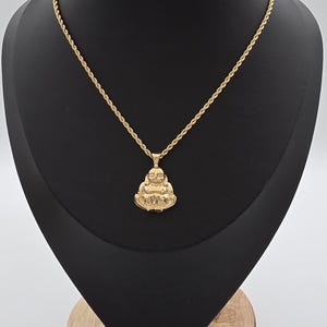 May include: A gold-toned necklace featuring a rope chain and a pendant shaped like a seated Buddha figure. The pendant has a polished finish and depicts the figure with hands resting on the lap. The chain is thin and textured with a twisted rope design.