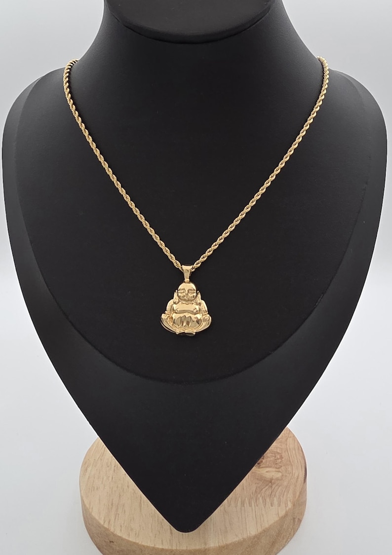 May include: A gold-toned necklace featuring a rope chain and a pendant shaped like a seated Buddha figure. The pendant has a polished finish and depicts the figure with hands resting on the lap. The chain is thin and textured with a twisted rope design.