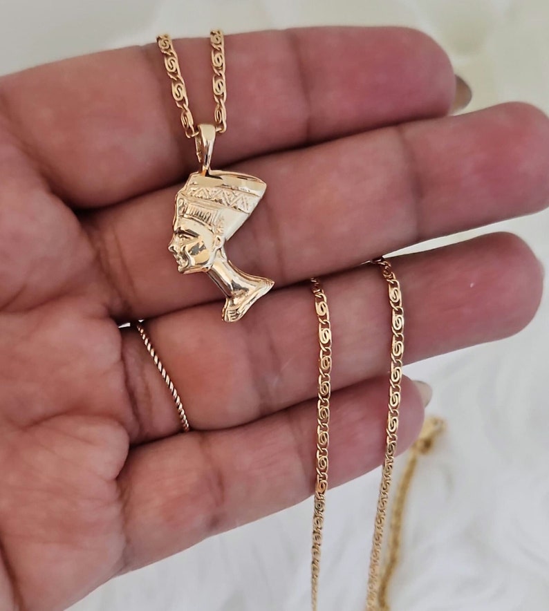 May include: Gold chain necklace with a pendant featuring a profile of an Egyptian queen wearing a headdress.